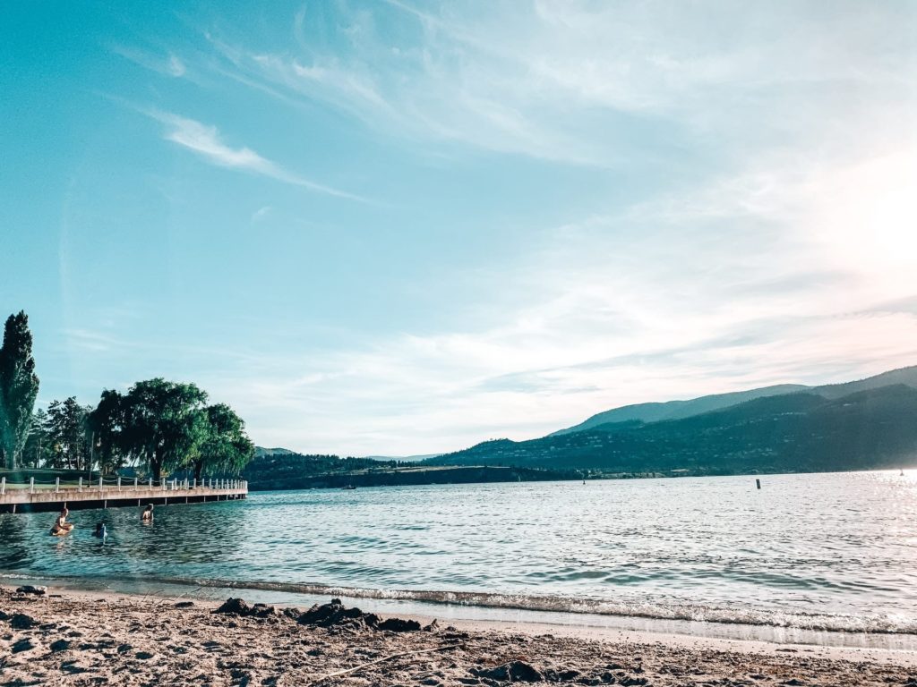 Okanagan lake beach with valley mountains in background on a clear sunny day