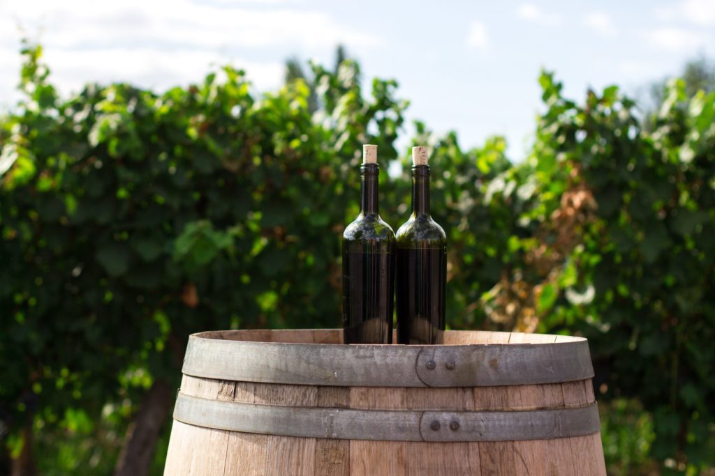 Two wine bottles on a wine barrel in a vineyard pertaining to Kelowna wineries and wine tastings for business professionals.